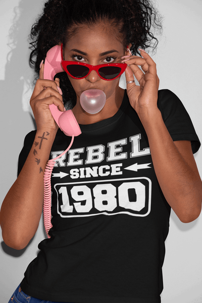 Woman Holding Retro pink phone, red frame sun glasses blowing gum wearing a black t-shirt with "Rebel since 1980" designed on front