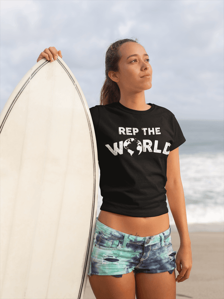 Surfer girl, standing beside surfboard wearing a black t-shirt with Rep the world design from the Xpert apparel store...